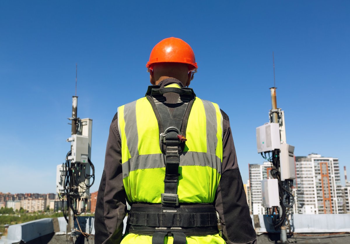Rooftop wireless technician in hardhat, hi-vis vest, and full safety harness working on cellular antennas