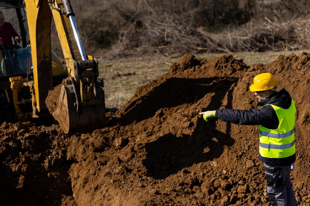 Construction worker in hardhat, hi-vis vest, safety glasses, and gloves signaling an excavator during earthwork