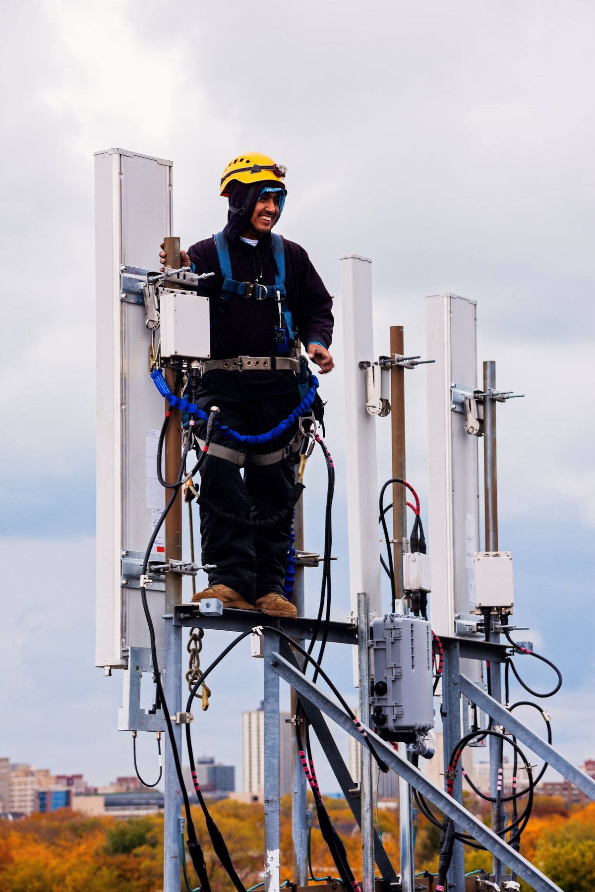 Tower crew leader in hardhat, safety glasses, gloves, and full harness working at the top of a cellular antenna array