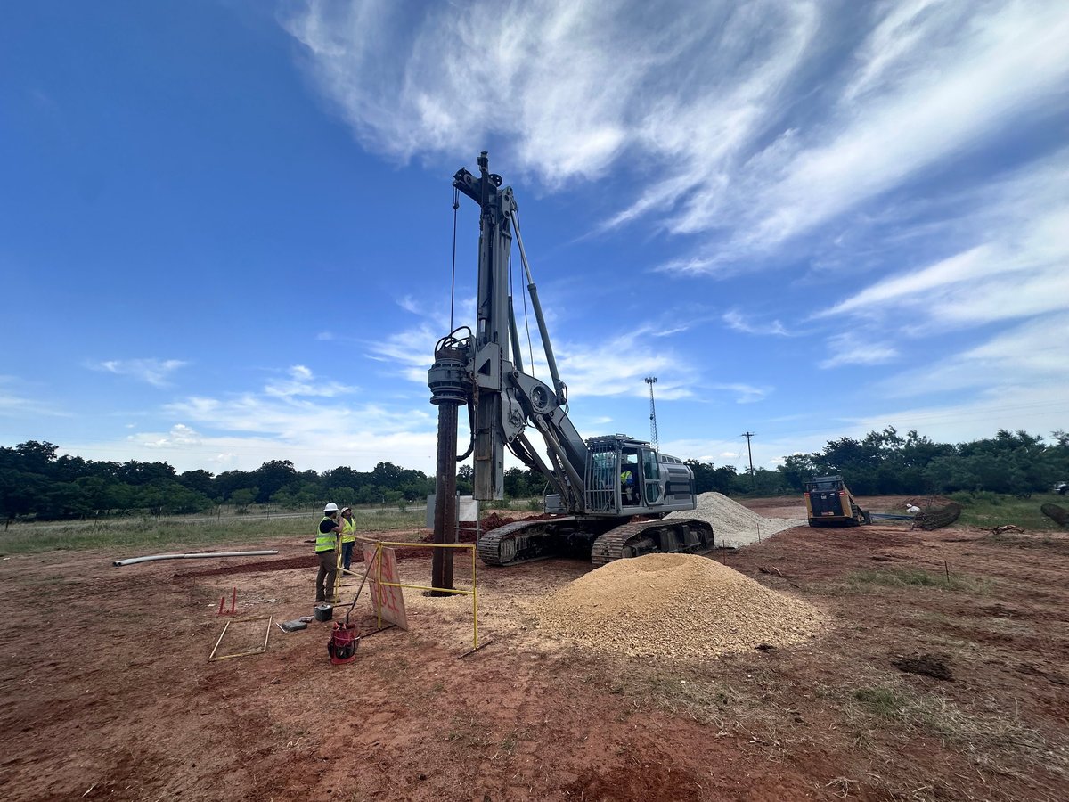 Large drilling rig on construction site with worker in safety gear, pile of excavated soil, clear blue sky