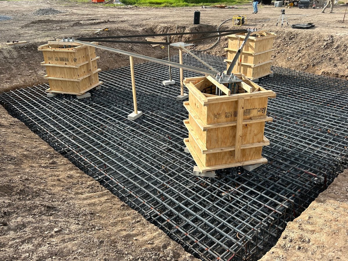 Construction site with black reinforced plastic mat, wooden support structures, and metal vertical posts during foundation preparation