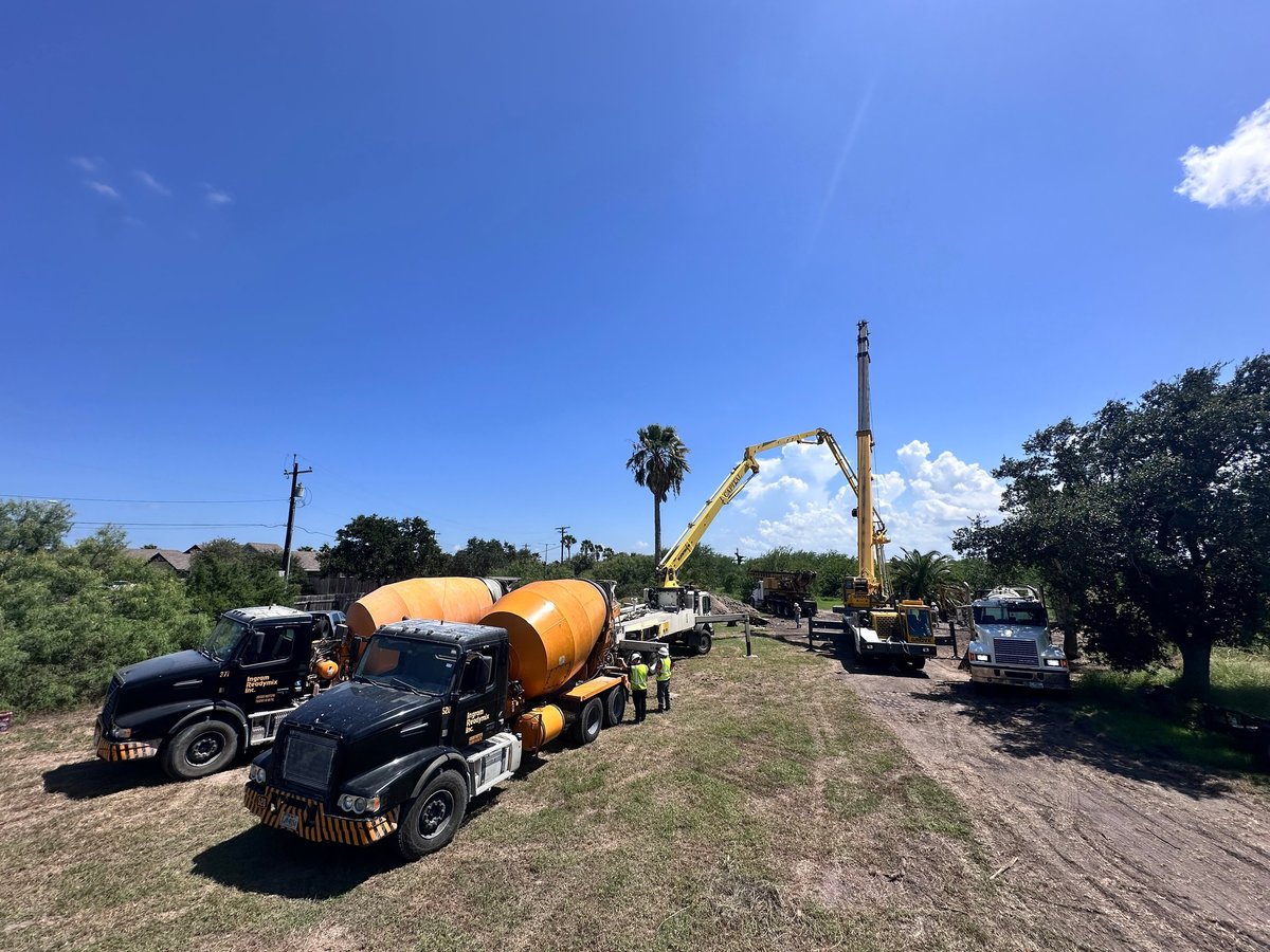 Heavy construction equipment including a concrete mixer truck and excavator parked on a rural dirt road under clear blue sky.